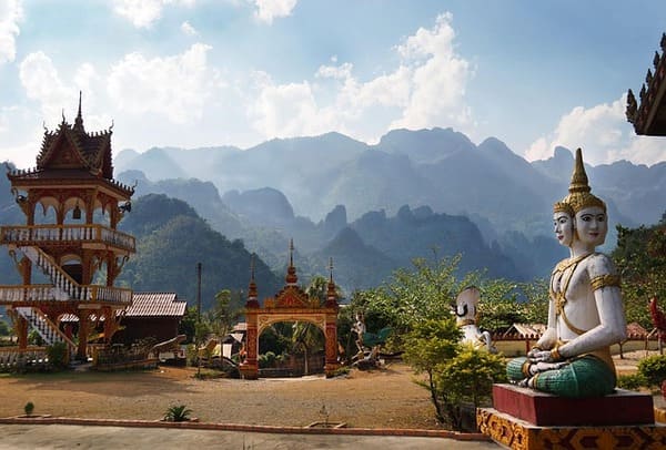 Temple bouddhiste traditionnel au Laos avec statues et montagnes en arrière-plan, un décor spirituel et naturel incontournable lors d’un voyage au Laos - voyage sur mesure avec Nahra Travel, spécialiste Asie et Moyen-Orient.